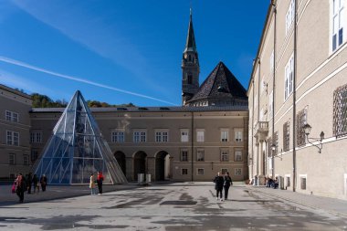 Salzburg, Avusturya - 01 Kasım 2023: Salzburg, Avusturya 'da arka planda bir kilise kulesi ve bir bina bulunan bir avluda cam bir piramit duruyor.