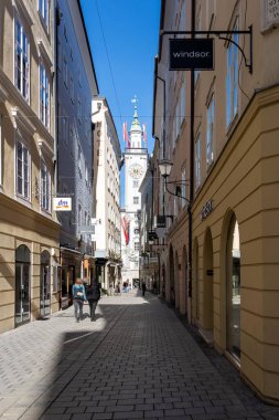 Salzburg, Avusturya - 01 Kasım 2023: Salzburg, Avusturya 'da dar bir cadde, uzaktaki bir saat kulesine giden binalar ve kaldırım taşlarıyla kaplı..