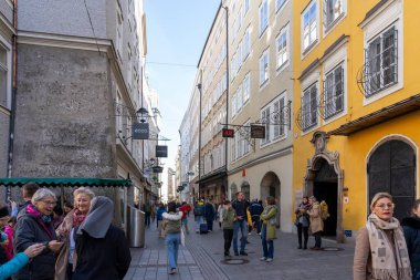 Salzburg, Avusturya - 01 Kasım 2023: Avusturya 'nın Salzburg şehrinde her iki tarafında da yüksek binalar bulunan dar bir cadde, dükkanların ve kafelerin önünden geçen insanlarla dolu.