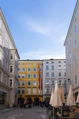 Salzburg, Avusturya - 01 Kasım 2023: Salzburg, Avusturya 'da her iki tarafında binaları olan dar bir kaldırım taşı caddesi. Sarı bir binada büyük bir Avusturya bayrağı sallanıyor. Birkaç kişi bir mağazanın önünde duruyor..