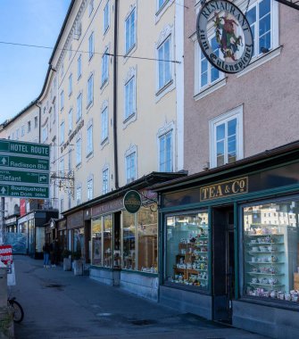 Salzburg, Avusturya - 01 Kasım 2023: Salzburg, Avusturya 'da mağazalarla dolu dar bir cadde. Binaların ön cepheleri ve geniş pencereleri var. Renkli kupalarla dolu bir çay dükkanı sokağın sonunda duruyor..