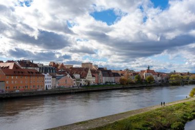 Regensburg, Almanya - 06 Kasım 2023: Regensburg, Almanya, taştan yolu ve yemyeşil çimenleri olan bir nehir sergiliyor. Şehirdeki gökyüzü bulutlu. Uzakta gökyüzü var..