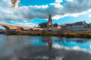 Regensburg, Almanya - 06 Kasım 2023: Almanya 'nın Regensburg kentinde tarihi binalar ve arka planda bir kilise kulesi bulunan bir taş köprü uzanır..