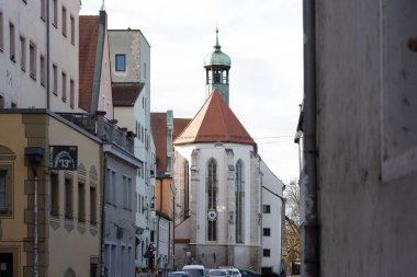 Regensburg, Almanya - 06 Kasım 2023: Almanya 'nın Regensburg şehrinde tarihi binalarla dolu dar bir cadde. Uzun çan kulesi olan bir kilise sahneyi yönetiyor..