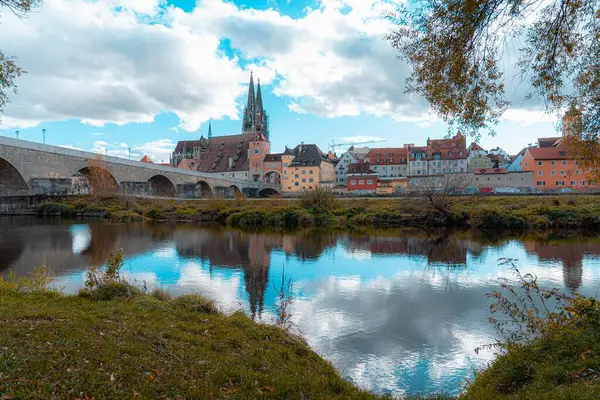 Regensburg, Almanya - 06 Kasım 2023: Almanya 'nın Regensburg kentinde bir nehir boyunca uzanan taş köprü, kıyılarda renkli binalar ve arka planda yüksek bir kilise bulunur..