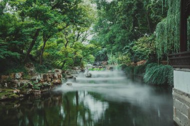 Durgun bir göletten yükselen sisli, yemyeşil ve taş bir köprüyle çevrili sakin bir Çin bahçesi..