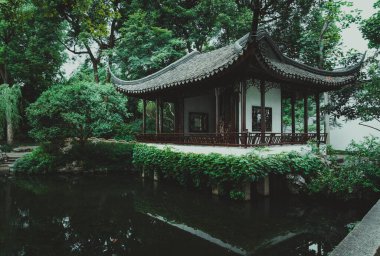 Eğri çatılı geleneksel bir Çin pavyonu durgun bir göletin üzerinde duruyor, yemyeşil çimlerle çevrili..