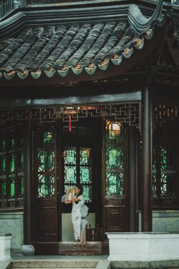 Suzhou, Çin - 11 Haziran 2024: Beyaz elbiseli bir kadın geleneksel bir Çin binasının kapısında duruyor. Giriş ahşap oymalar ve karmaşık kafes işçiliğiyle çerçevelenmiş..
