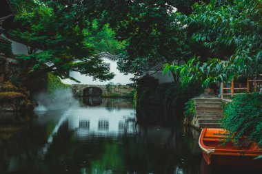 Suzhou bahçesinde sakin bir gölet. Su durgun ve çevresindeki yeşilliği ve beyaz yapıyı yansıtıyor..