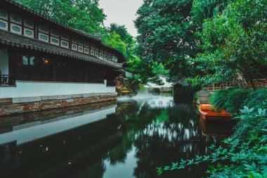 Solunda geleneksel bir bina ve sağında küçük bir tekne bulunan Çin bahçesindeki sakin bir kanal yemyeşil alanda esiyor..