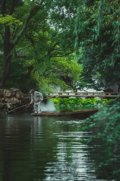 stock image Suzhou, China - June 11, 2024 : A man rows a boat through a tranquil pond, surrounded by lush greenery and a stone bridge in the Humble Administrator's Garden in Suzhou, China.