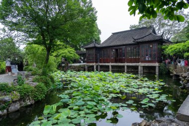 Suzhou, Çin - 11 Haziran 2024: Ahşap pavyonu, yemyeşil yaprakları ve taş yolları olan bir Çin bahçesinde bir gölet.