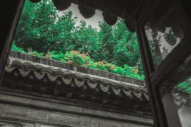 Eski bir Çin binasının önündeki yemyeşil bir bahçeye bakan pencereden bir manzara. Binanın gri fayanslı bir çatısı ve karmaşık bir şekilde oyulmuş taş bir duvarı var..