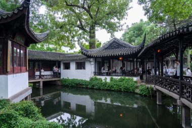 Suzhou, Çin - 11 Haziran 2024: Eğri çatıları ve beyaz duvarları olan geleneksel Çin mimarisi. Tahta bir köprü çevredeki binaları ve yeşillikleri yansıtan bir göleti geçer..