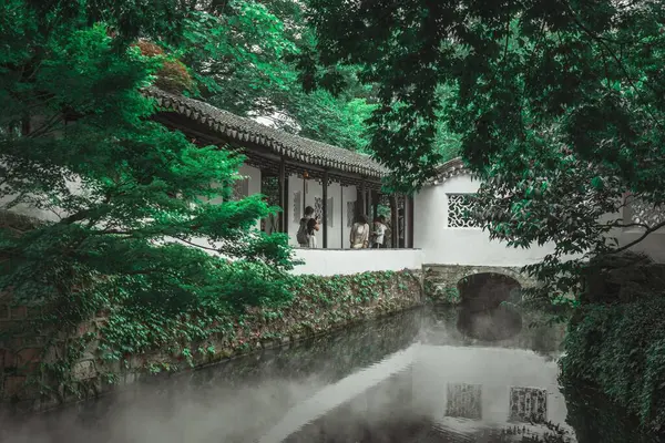 stock image Suzhou, China - June 11, 2024 : A tranquil canal winds through a lush garden, with mist rising from the water and green foliage framing the scene.