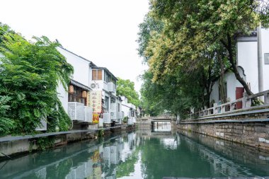 Suzhou, Çin - 11 Haziran 2024: Geleneksel bir Çin kasabasının kalbinden geçen dar bir kanal. Taş duvarlar suyun kenarında, yemyeşil ve canlı yansımalarla süslenmiş..