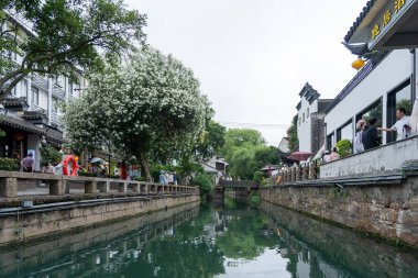 Suzhou, Çin - 11 Haziran 2024: Geleneksel bir Çin kasabasından dar bir kanal akar. Yanları ağaçlar ve yeşilliklerle kaplı binalar doğanın bir dokunuşunu ekliyor. İnsanlar yolda yürüyor, manzaranın tadını çıkarıyorlar..