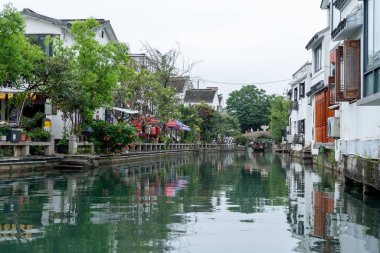 Suzhou, Çin - 11 Haziran 2024: Beyaz binalar, yeşillikler ve taş bir köprü ile geleneksel bir Çin kasabasından geçen bir kanal..
