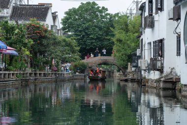 Suzhou, Çin - 11 Haziran 2024: Küçük bir tekne Çin 'in Suzhou kentindeki dar bir kanal üzerinde taş bir köprünün altında süzülüyor. Kanal boyunca uzanan binalar, beyaz duvarları ve fayanslı çatıları olan geleneksel Çin mimarisidir. Kanalı çevreleyen ağaçlar...