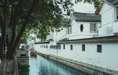 Dar bir kanal geleneksel bir Çin kasabasından geçiyor, su kenarında beyaz binalar var..