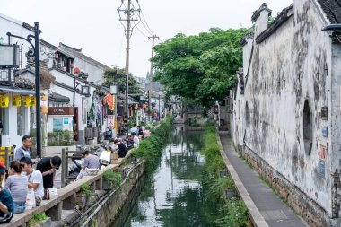 Suzhou, Çin - 11 Haziran 2024: Geleneksel Çin binaları ve yürüyüş yolundaki insanlarla dolu bir Suzhou mahallesinden bir kanal geçiyor.