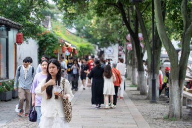 Suzhou, Çin - 11 Haziran 2024: Kalabalık bir Asya şehrinde ağaçlarla kaplı bir caddede yürüyen genç bir kadın.