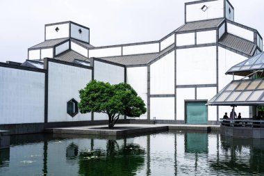 Suzhou, Çin - 12 Haziran 2024: Önünde büyük bir gölet bulunan modern bir bina. Bina siyah aksanlı beyaz ve eşsiz bir geometrik tasarımı var. Gölette su zambakları ve binanın bir yansıması var..