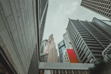 Düşük açılı, Hong Kong 'da yükselen gökdelenlerin yakın çekim görüntüsü, gri ve bej cepheleri sessiz bir gökyüzüne doğru yükseliyor. Şehrin yoğun mimarisi dramatik ve yükselen bir perspektif yaratıyor..