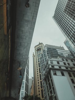 Bir Hong Kong yakın çekimi, yüksek gökdelenleri gözler önüne seriyor, dış cepheleri, çağdaş taş ve modern camdan oluşan bir karışım, sessiz bir gökyüzüne doğru uzanıyor. Perspektif aşağıdan, binaların uçsuz bucaksız yüksekliğini vurguluyor.