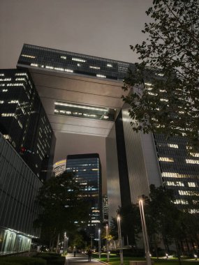 Hong Kong, Çin - 12 Mart 2025: Aydınlatılmış pencereli, uzun ve modern bir binanın gece görüşü. Ağaçlar ve patikalar ön planda karanlık, bulutlu bir gökyüzünün altında..