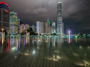 Hong Kong, Çin - 12 Mart 2025: Yüksek binaların aydınlatıldığı ve suya yansıtılan gece şehir manzarası. Şehir ışıkları karanlık gökyüzünü aydınlatır ve canlı bir sahne yaratır..