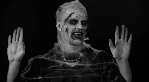 Zombie man with makeup with wounds scars and white contact lenses wearing earphones, listening music, dancing, celebrating on black background. Sinister dead guy. Halloween, filming, staging concept
