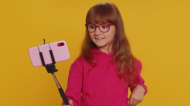 Genç kız çocuk blogcu kız akıllı telefon özçekim çubuğuyla selfie çekiyor abonelerle internet üzerinden video görüşmesi yapıyor. Stüdyo sarı arka planda izole edilmiş küçük çocuklar.