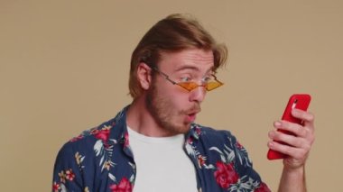 Akıllı telefon görünümlü mutlu turist samimi bir sevinç gösterisi kazanır, para dolar banknotları alır, başarı piyangosunda şans getirir. Genç sakallı çocuk bej renkli stüdyoda tek başına izole edilmiş.