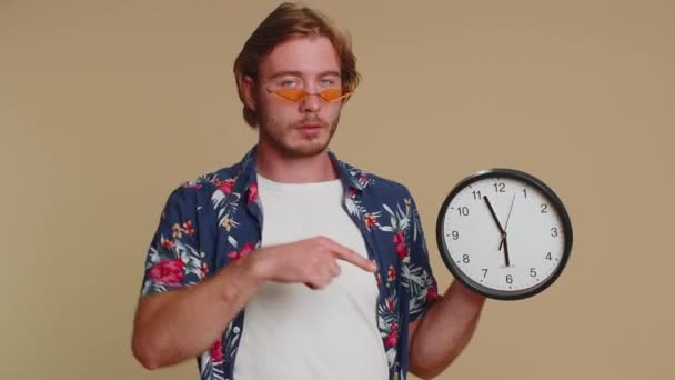 这是你的时间 穿着衬衫 留着胡子的年轻人的画像 在钟表上显示时间 竖起大拇指 用手指对着相机 在室内拍摄的一个成年人在米色背景下被单独拍摄 — 图库视频影像