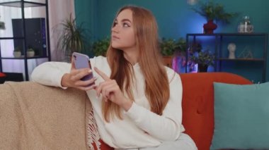 Portrait of adult girl sitting on sofa uses mobile phone smile at modern home apartment. Young woman texting share messages content on smartphone social media applications online, watching relax movie