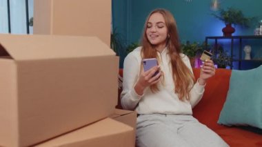 Alışveriş manyağı kız elinde hediye karton kutusuyla evde oturmuş online sipariş veriyor, kredi kartı ile ödeme yapıyor. Evdeki kanepede oturan genç bir kadın. İnternette indirimli satış