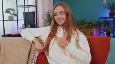 Like. Happy lovely adult girl looking approvingly at camera showing thumbs up, like sign positive something, good news, positive feedback. Young woman sitting on orange couch at home living room