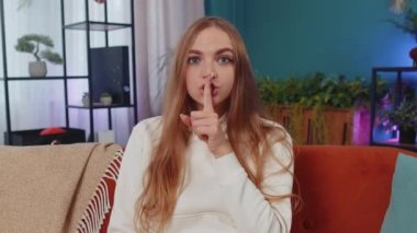 Shh be quiet please. Adult caucasian girl presses index finger to lips makes silence hush gesture sign do not tells gossip secret. Young woman at home apartment living room sitting on orange couch