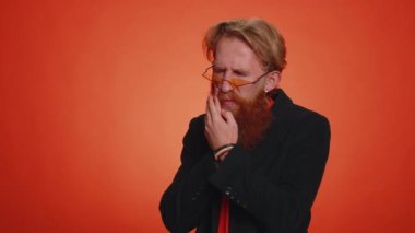 Diş ağrısı ya da diş eti iltihabından muzdarip yaralı yanağa dokunan sakallı genç bir adam dişçi randevusu ya da diş eti hastalığı bekliyor. Kapalı alanda çekilen yetişkin adam turuncu arka planda tek başına.