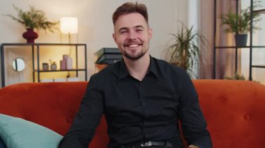 Gibi. Mutlu, sevimli yetişkin bir adam onaylayıcı bir şekilde kameraya bakıyor olumlu bir şey imzalıyor, iyi haber, olumlu geri bildirim gibi. Oturma odasındaki turuncu koltukta oturan sakallı genç adam.