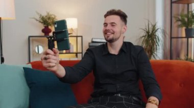 Yakışıklı erkek blogcu cep telefonu kamerasından selfie çeken cihaz ekranına bakıyor, modern sosyal medya vlog uygulamasında sanal video görüşmesi yapıyor. Evdeki koltukta akıllı telefonu olan adam.
