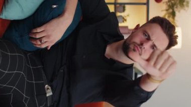 Hoşlanmamak. Sinirli adam işaret dilini gösteriyor, hoşnutsuzluğunu dile getiriyor, hoşnutsuzluğunu dile getiriyor, modern ev dairelerinde tatminsiz kötü işler yapıyor. Kanepede oturan hoşnutsuz genç adam. Dikey görünüm