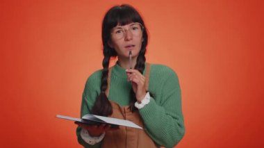 Düşünceli, yeşil kazaklı, not defteri günlüğüne kalemle düşünceler yazan bir gazeteci kadın, liste yapmak için, iyi fikir. Portakal rengi stüdyoda izole edilmiş genç bir kız.