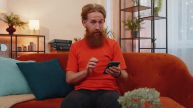 Portrait of man use mobile smartphone typing browsing say Wow yes found out great big win good news celebrate lottery jackpot doing winner gesture. Young guy at home in living room sitting on sofa