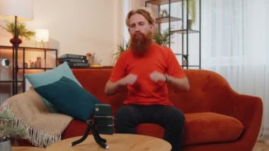 Beyaz sakallı adam blogcusunun portresi akıllı telefon tripoyla selfie çekiyor, dans ediyor, video blogu kaydediyor, abonelerle çevrimiçi iletişim kuruyor. Evdeki genç adam turuncu koltukta oturuyor.