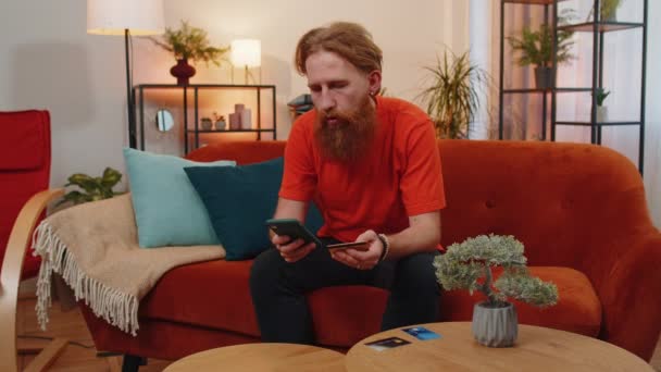 有胡子的男子在汇款 网上购物 在家里订购食物时使用信用卡和智能手机 客厅里坐在沙发上的红头发小伙子 — 图库视频影像