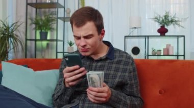 Aile bütçesi planlamak. Gülümseyen genç adam nakit para sayıyor, akıllı telefon kullanıyor evdeki faturaları hesaplıyor. Neşeli genç adam gelirinden memnun ve planlı tatil ve hediyeler için para biriktiriyor.