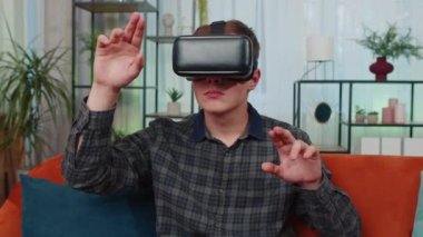 Genç adam evde VR gözlük kullanıyor, 360 boyutlu video konseptinin tadını çıkarıyor, eğleniyor, olumlu ruh halinde, ellerini havada hareket ettiriyor, oturma odasında kanepede oturuyor. Sanal gerçeklik, modern aygıt