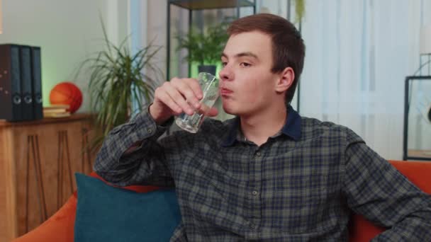 口渴的少年男子坐在室内拿着天然水杯子的画像 会让他们喝着静水 防止脱水 有良好生活习惯 健康体形 体重减轻观念的人 — 图库视频影像
